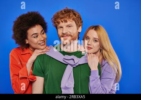Alternative Beziehungen, Polyamorien, Porträt von drei Personen, junger rothaariger Mann und wunderschöne multirassische Frauen auf blauem Hintergrund, Studioaufnahme, Vib Stockfoto