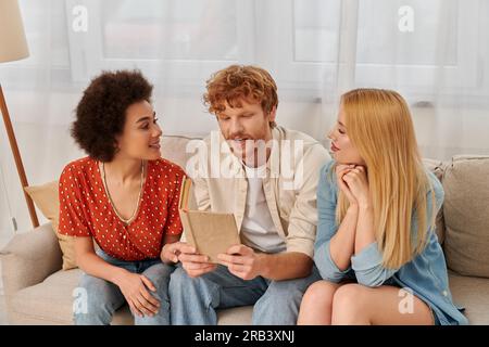 Offene Beziehungen, rothaariger Mann liest Buch für multikulturelle Frauen im Wohnzimmer, nicht traditionelle Liebhaber, kulturelle Vielfalt, Romantik und Liebe, Mode Stockfoto