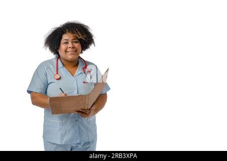 Junge afrolateinische Ärztin, die eine blaue Uniform und ein rotes Stethoskop am Hals trägt, lächelt in die Kamera und hält ein offenes braunes Stockfoto