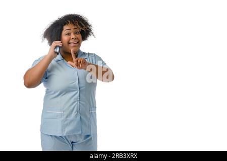 Junge afrolateinische Ärztin in blauer Uniform steht am Telefon lächelnd und spricht mit einem erhobenen Finger und einem Frageausdruck auf ihr Stockfoto