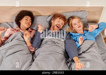 Polyamorien-Konzept, drei Erwachsene, Männer und Frauen aus verschiedenen Rassen in Pyjamas, die zusammen aufwachen, morgens, unter der Decke, Schlafzimmer, kulturelle Vielfalt, Bisexua Stockfoto