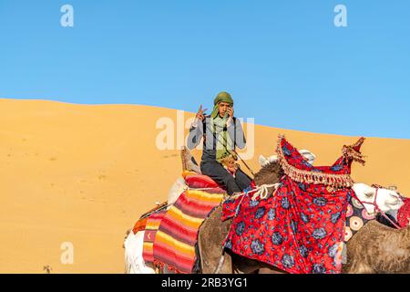 Saoura, Algerien - 28. Dezember 2022: Ein junger tuareg-Mann reitet auf einem weißen Dromedärkamel, das mit einem farbigen Stoffsattel verziert ist und mit zwei Fingern posiert. Stockfoto