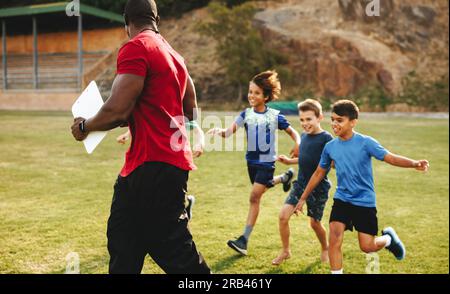 Sporttrainer wird von seiner Sportmannschaft verfolgt. Schulsportmannschaft läuft zu ihrem Trainer. Coach und Schüler spielen spielerisch auf dem Sportplatz. Stockfoto