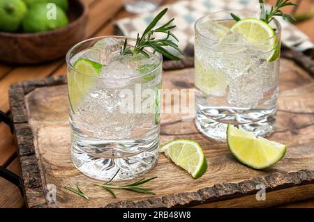 Gin- und Tonic-Cocktail garniert mit Limettenkeilen und Rosmarin auf rustikalem Holztablett Stockfoto