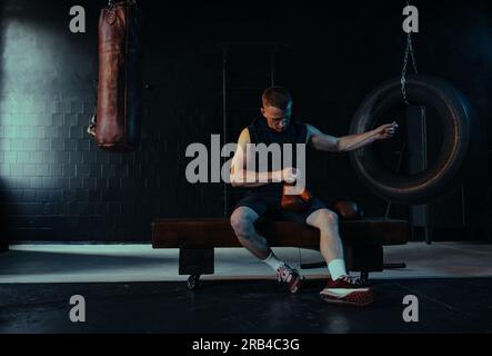 Männlicher Boxer sitzt auf einer Bank in einem dunklen Fitnessstudio und zieht Boxhandschuhe an, um vor dem Hintergrund einer Stanztasche zu trainieren. Stockfoto