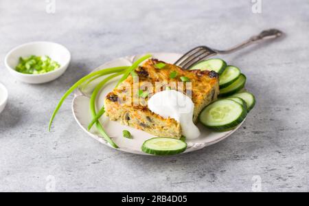 Gemüseauflauf, kugel, aus Kartoffeln, Karotten und Pilzen mit Sauerrahm, Gurkenscheiben und grünen Zwiebeln auf grauem, strukturiertem Hintergrund Stockfoto