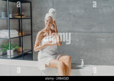 Die träumige europäische Frau trägt Körpercreme, eingewickelt in ein Handtuch. Gesundheit, Beauty-Konzept. Badezimmerverwöhnung. Stockfoto