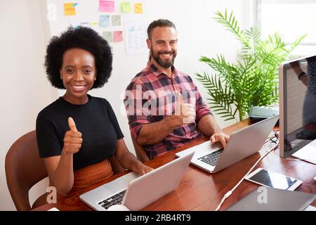 Zwei Entwickler, die an einem gemeinsam genutzten Schreibtisch sitzen, geben Daumen nach oben und programmieren im Büro auf einem Laptop Stockfoto