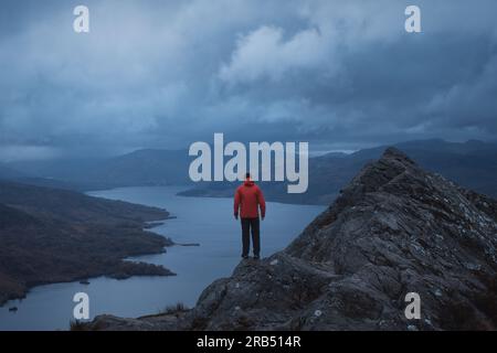 Ein Wanderer in einer roten Jacke auf einem Berg blickt auf die herrliche Landschaft eines Sees, der von Bergen umgeben ist. Loch Katrine. Loch Lomond und der Trossachs-Nationalpark. Schottland Stockfoto