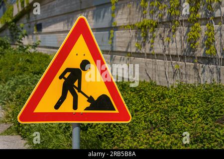 Warnschild für Straßenarbeiten mit grünen Büschen und Betonwand im Hintergrund Stockfoto