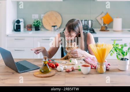 Die Katze und ihr Besitzer in Schürzen kochen gemeinsam in der heimischen Küche. Hauskatze beobachtet, wie das Mädchen das Futter salzt. Stockfoto