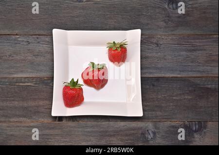 Drei Erdbeeren auf einem wunderschönen weißen Teller auf einem Holzhintergrund. Stockfoto