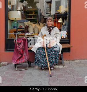 Eine alte Dame mit Hijab sitzt vor einem Geschäft in Istanbul auf einer Bank und hält einen Gehstock mit ihrem Einkaufswagen neben sich, Truthahn. Stockfoto