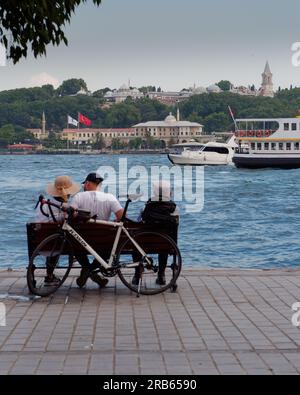 Ein Paar und ein älterer Mann sitzen an einem Sommerabend auf einer Bank in Karakoy mit Blick auf das Goldene Horn und den Topkapi-Palast, Istanbul, Türkei Stockfoto
