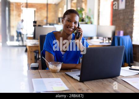Portrait einer glücklichen afroamerikanischen Geschäftsfrau, die sich auf dem Smartphone unterhält und zu Mittag isst. Geschäft, Lebensmittel, Technologie und Arbeit, unverändert. Stockfoto