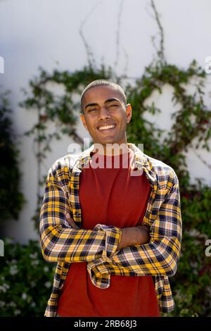 Porträt eines glücklichen birassischen Mannes, der mit gekreuzten Armen steht und im sonnigen Garten lächelt. Sommer, Freizeit und Lebensstil, unverändert. Stockfoto