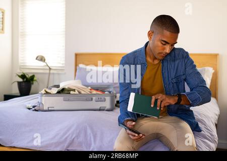 Ein birassistischer Mann mit Pass und Smartphone, der auf dem Bett mit dem Koffer sitzt und die Uhr überprüft. Reisen, Urlaub, Vorbereitung, Zeit, Kommunikation, Kostenlose t Stockfoto