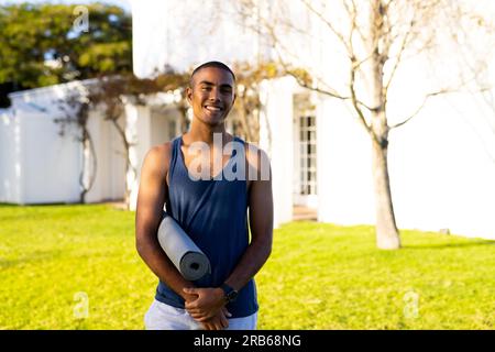 Porträt eines glücklichen birassischen Mannes, der Yoga-Matte im sonnigen Garten hält. Sommer, Fitness, Wohlbefinden und gesunder Lebensstil, unverändert. Stockfoto