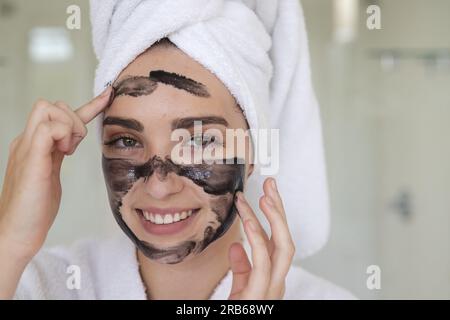 Porträt einer glücklichen Weißen mit Handtuch auf dem Kopf, die Maske auf dem Gesicht im Badezimmer macht, Kopierbereich. Häusliches Leben, Selbstfürsorge und Lebensstil, unverändert. Stockfoto