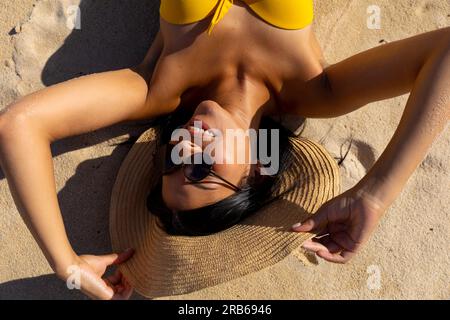 Blick von oben auf eine glückliche, birassische Frau mit Sonnenhut und Sonnenbrille, die auf Sand am sonnigen Strand liegt und lächelt. Sonnenbaden, Sommer, Freizeit, Entspannung, Lifest Stockfoto