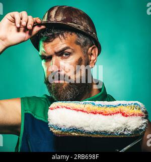 Seriöser Maler oder Dekorateur in Schutzhelm mit Malerwalze. Handwerker, Handwerker, Handwerker im Schutzhelm mit Malerwalze. Professioneller Maler Stockfoto