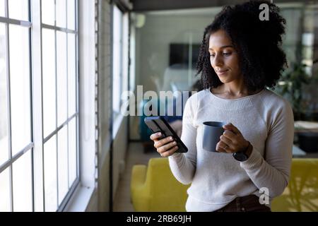 Glückliche, birassistische, legere Geschäftsfrau mit Kaffee, die ihr Smartphone benutzt, steht im Büro, Kopierbereich. Arbeit, Arbeit, Kommunikation und ungezwungenes Büro, unal Stockfoto