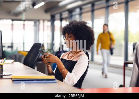 afroamerikanische Geschäftsfrau, die am Schreibtisch sitzt und im Büro ein Smartphone benutzt. Ungezwungene Geschäfte, Kommunikation und Arbeit, unverändert. Stockfoto