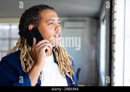 Birassistischer, lässiger Geschäftsmann mit Dreadlocks, der auf dem Smartphone spricht und am Fenster in einem sonnigen Büro steht. Arbeit, Arbeit, Kommunikation und ungezwungenes Büro, Stockfoto