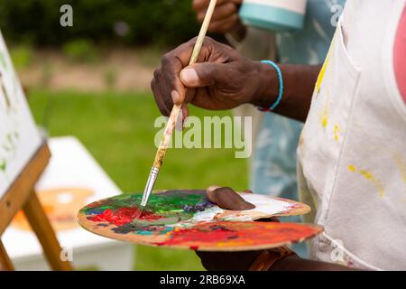 Mitten in der afroamerikaner-Malerei an der Staffelei und Kaffee im Garten. Seniorenleben, Ruhestand, Zusammensein, Kreativität und Stockfoto