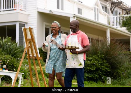 Glückliches Senior-afroamerikanisches Paar, das an der Staffelei malt und Kaffee im Garten trinkt. Lebensstil für Senioren, Ruhestand, Zusammensein, Kreativität und Hobbys Stockfoto