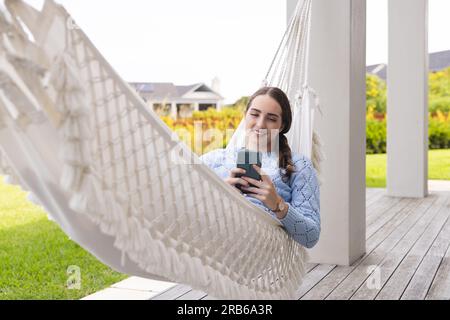 Glückliche weiße Frau, die in einer Hängematte lag und ihr Smartphone im Garten benutzte. Häusliches Leben, Technologie und Lebensstil, unverändert. Stockfoto