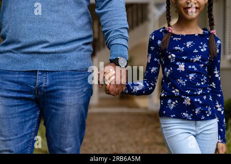 Mittelteil eines birassischen Vaters und einer lächelnden Tochter, die im Garten vor dem Haus Händchen hält. Familie, Liebe, Zusammensein, Heim, Lebensstil und häusliche Gesundheit Stockfoto