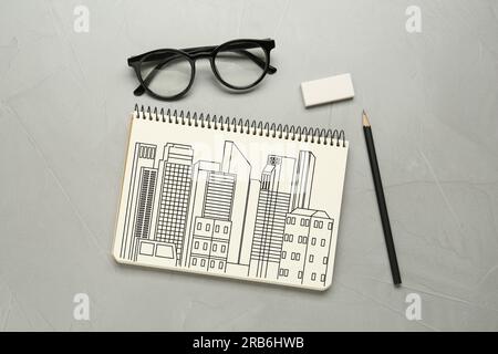 Skizze von Gebäuden in Notizbuch, Bleistift, Radiergummi und Brille auf grauem Tisch, flacher Unterlage Stockfoto
