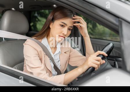 Eine müde junge Asiatin mit starken Kopfschmerzen sitzt in ihrem geparkten Auto hinter dem Steuer und schnallt sich an, während sie ihre Hand über die Stirn hält Stockfoto