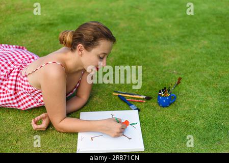 Junge Künstlerin und Illustratorin, die einen Bleistift zieht und im Sommer auf dem Rasen liegt Stockfoto