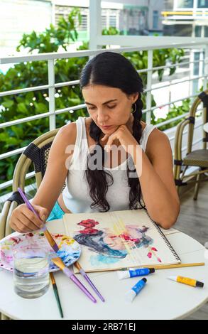 Eine braune ukrainische Frau malte Aquarell eines asiatischen Mädchens mit einem Pinsel auf einem Tisch mit Wasser und Farben Stockfoto