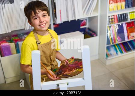 Attraktiver lächelnder Teenager, talentierter kleiner Künstler, der eine Palette mit Ölfarben in der Hand hält, an einer Staffelei vor dem Hintergrund der Leinwand gemalt wird. Ar Stockfoto