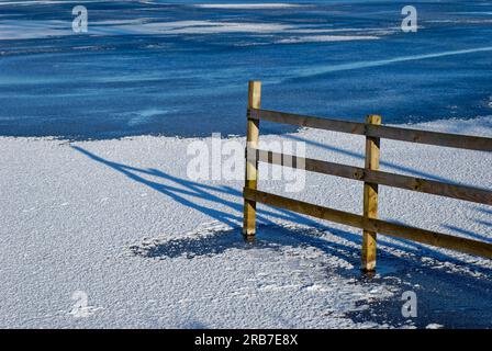 Ein Holzzaun am Rande des Murton Loch mit der Wintersonne, die lange Schatten über dem gefrorenen Wasser wirft. Stockfoto