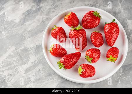 Reife Gartenerdbeere auf weißer Platte auf grauem Hintergrund. Frisch gepflückte Erdbeeren auf Beton. Speicherplatz kopieren. Draufsicht Stockfoto