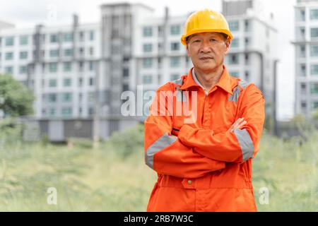 Seriöser und entschlossener asiatischer Ingenieur mittleren Alters in orangefarbenem Overall mit Schutzhelm, der seine Arme kreuzte, während er stolz vor seinem Multi-Stori stand Stockfoto
