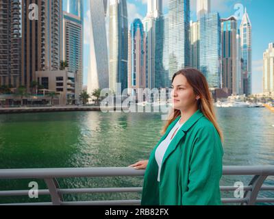 Junge Frau im grünen Anzug geht durch die Straßen von Dubai, Dubai Marina District. Reisekonzept der Vereinigten Arabischen Emirate. Die Vorstellung von erfolgreichem Auswanderer, Umzug in ein anderes Land, Arbeitsvisum Stockfoto