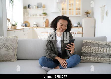 Fröhliches Mädchen chattet online, nutzt Handy-Apps, sitzt zu Hause auf dem Sofa. Gemischte Rassen Ladys online einkaufen, lächeln. Stockfoto