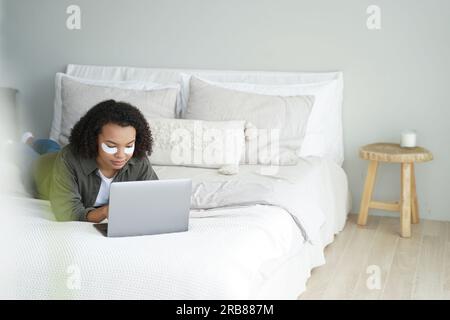 Afroamerikanischer Teenager mit Augenklappen funktioniert auf einem Laptop im Bett. Birassistische Frauen kaufen online während der morgendlichen Hautpflege. Stockfoto