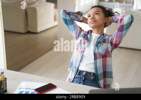 Ein träumiger, birassistischer Teenager macht eine Pause vom Lernen zu Hause, lehnt sich mit den Händen hinter dem Kopf zurück, in Gedanken verloren. Stockfoto