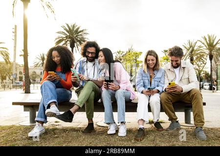 Multiethnische Gruppe von Freunden, die auf ihren Smartphones auf einer Bank auf der Straße SMS schreiben. Verschiedene junge Leute lächeln und benutzen ihr Telefon Stockfoto