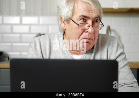 Ein grauhaariger Mann mit Brille, der am Laptop sitzt und ein Fischgesicht mit Lippen macht. Verrückte und komische Geste. Lustiger alter Großvater mit verspieltem Stockfoto