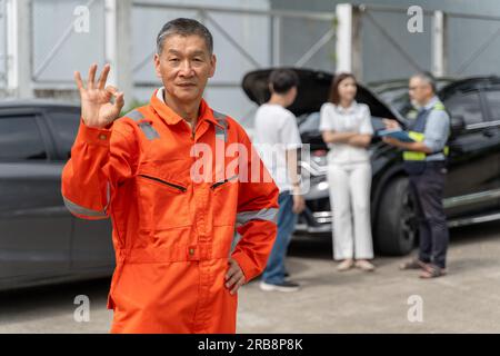 Ein männlicher Autoverkäufer im orangefarbenen Overall, der stolz auf sich steht und seine rechte Hand mit einem OK-Zeichen hochlegt und seine linke Hand auf der Hüfte, ein anderes Alter Stockfoto