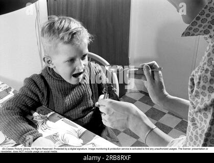 Ein Junge ist krank und nimmt seine Medizin nicht. Seine Mutter hält einen Löffel und wartet darauf, dass er ihn nimmt, und das tut er. Er öffnet seinen Mund und nimmt ihn, obwohl er nicht so gut schmeckt. Schweden 1956. Conard Ref. 3157. Stockfoto