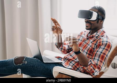 Lächelnder afroamerikanischer Mann mit Virtual-Reality-Brille, der im Cyberspace arbeitet oder Videospiele auf einem Laptop spielt, sitzt im Stuhl. Modernes schwarzes Mal Stockfoto