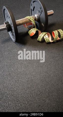 Schwarze, rostige, ladbare Hantel und Maßband auf dem Boden im Fitnessstudio. Schwarzer Hintergrund und leerer Platz für Text. Fitness, Krafttraining Stockfoto